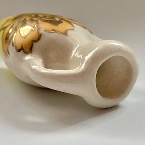 Puede incluir: Una pequeña jarra de cerámica blanca con un diseño floral dorado. La jarra tiene un asa y una abertura redonda.