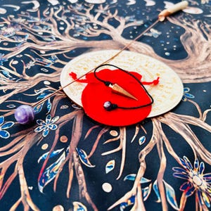 Pode incluir: Um tabuleiro de pêndulo de madeira com um pêndulo dourado e uma bolsa vermelha contendo um pêndulo de ponta de cristal. O tabuleiro tem um design de árvore da vida com fundo preto e detalhes dourados. O tabuleiro apresenta letras, números e símbolos.