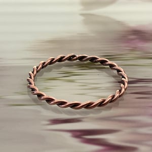 May include: A copper-colored ring with a twisted rope design. The ring is circular and sits on a blurred background with shades of pink and green. The ring's texture is visible, showcasing the intricate twists.