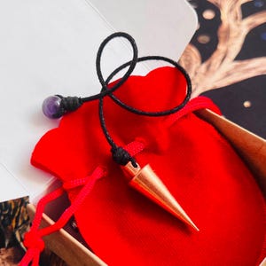 May include: A copper-colored metal pendulum with a pointed cone shape attached to a black cord. The cord features a small, round, purple amethyst bead near the end. The pendulum rests inside a bright red velvet drawstring pouch within a box.