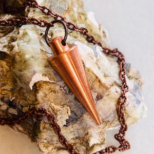 Op de afbeelding: Een koperkleurige pendelketting met een puntige, conische hanger. De hanger hangt aan een kleine ring en hangt aan een bijpassende koperen ketting. De ketting wordt gepresenteerd op een getextureerd, gebroken witte ondergrond.