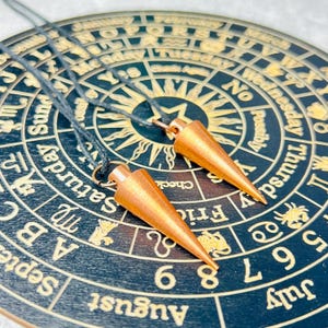 Può includere: Due pendoli da radiestesia di colore rame appesi a corde nere sopra un pannello circolare di legno con sfondo nero e lettere dorate. Il pannello presenta un calendario, i giorni della settimana e i simboli dello zodiaco.
