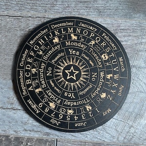 K&ouml;nnte beinhalten: Ein schwarzes, rundes Divinationsbrett aus Holz mit goldenen Buchstaben und Symbolen. Das Brett zeigt das Alphabet, Zahlen, Wochentage, Monate, Sternzeichen und die W&ouml;rter "Ja", "Nein" und "M&ouml;glicherweise".