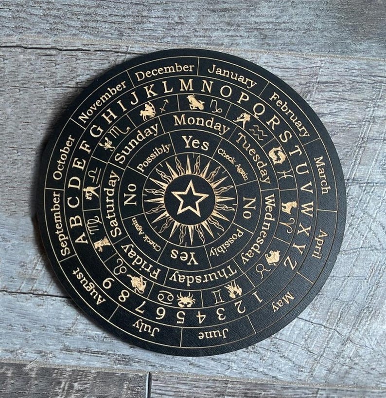 K&ouml;nnte beinhalten: Ein schwarzes, rundes Divinationsbrett aus Holz mit goldenen Buchstaben und Symbolen. Das Brett zeigt das Alphabet, Zahlen, Wochentage, Monate, Sternzeichen und die W&ouml;rter "Ja", "Nein" und "M&ouml;glicherweise".