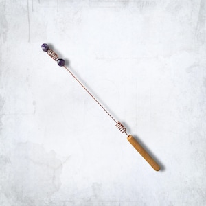 May include: A long, thin copper wire wand featuring a wooden handle at one end and a decorative section at the other. The top end includes two round, dark purple, mottled beads separated by a small copper wire coil, with another coil near the handle.