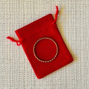 Könnte beinhalten: Ein dünner, kreisförmiger Ring mit einer gedrehten, seilartigen Textur aus einem metallischen, kupferfarbenen Material. Der Ring liegt auf einem kleinen, rechteckigen, leuchtend roten Stoffbeutel mit Kordelzug und passenden roten Bändern.
