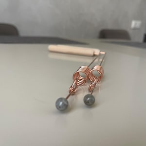 Op de afbeelding: Twee koperkleurige haarstyling tools met spiraalvormige ontwerpen en grijze kraalaccenten. Een houten handvat is op de achtergrond. De tools zijn ontworpen voor haarstyling en het creëren van krullen.
