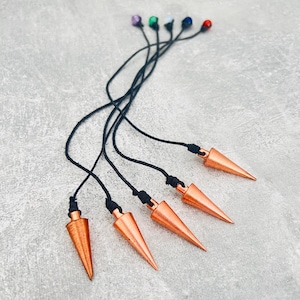 Pode incluir: Cinco pêndulos cor de cobre com pontas e cordões pretos. Cada pêndulo tem uma conta de cor diferente no topo do cordão: roxo, verde, branco, azul e vermelho. Os pêndulos estão dispostos sobre uma superfície cinza.