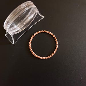 May include: A copper-colored, twisted wire ring sits on a black surface next to a clear plastic container. The ring is circular and appears to be handmade. The twisted design gives the ring a textured appearance.