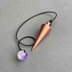 May include: A copper-toned metal pendulum with a smooth, conical shape attached to a thin black cord. At the end of the cord, there is a small, round, translucent purple bead with white marbling, providing a contrast to the metallic point.