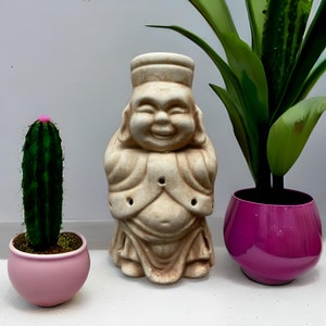 Könnte beinhalten: Eine weiße Keramikfigur eines lächelnden Buddha, der eine Robe trägt. Die Figur steht auf einem Tisch mit einem rosa Kaktus in einem Topf und einer grünen Pflanze in einem rosa Topf.