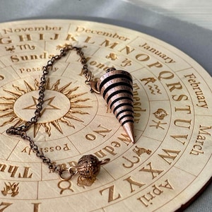 Könnte beinhalten: Ein kupferfarbenes Pendel mit Spiralmuster hängt über einem hölzernen Wahrsagebrett. Das Brett zeigt eingravierte Texte und Symbole, darunter Monate und astrologische Zeichen. Das Pendel ist an einer Kette befestigt und hat ein kugelförmiges Gewicht.