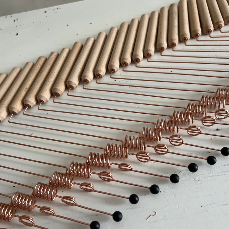 May include: A wooden wall hanging with multiple copper wire strands, each with a spiral and a black bead at the end. The wooden pieces are arranged in a staggered pattern, creating a wave-like effect.