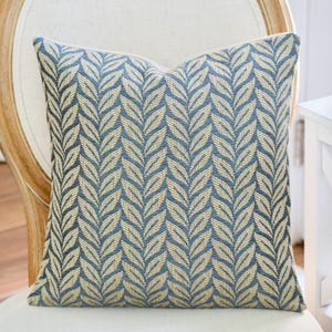 May include: Square decorative pillow with a blue and beige leaf pattern. The pillow is on a cream-colored chair. The pillow's design features a repeating pattern of stylized leaves in shades of blue and beige, creating a textured appearance.