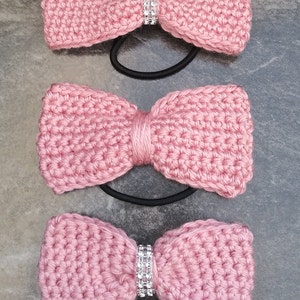 May include: Three pink crocheted hair bows with rhinestones. Each bow is attached to a black elastic hair tie.