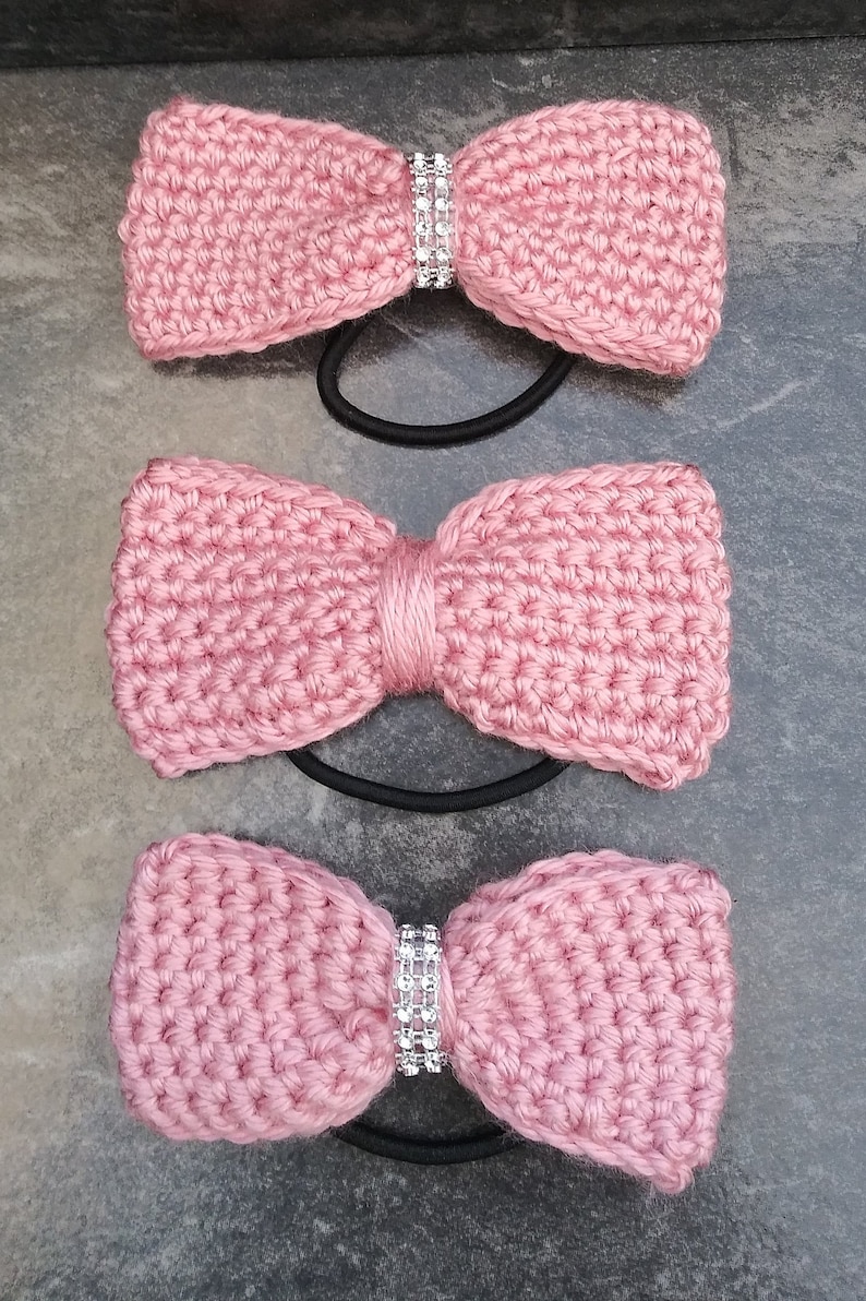 May include: Three pink crocheted hair bows with rhinestones. Each bow is attached to a black elastic hair tie.