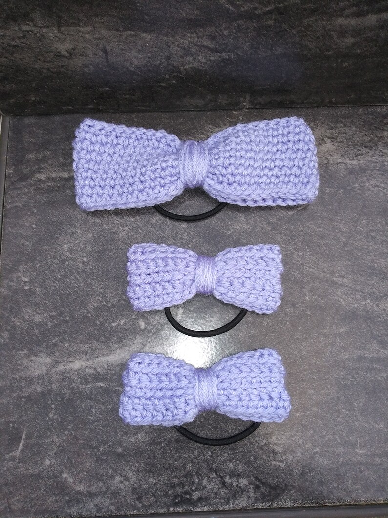 May include: Three purple knitted hair scrunchies in different sizes. The scrunchies are tied in a bow shape.