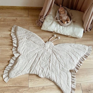 Könnte beinhalten: Eine beige, schmetterlingsförmige Spielmatte mit gerüschten Kanten. Die Matte besteht aus einem weichen, gesteppten Stoff und weist detaillierte Nähte auf, die an Schmetterlingsflügel erinnern. Eine Puppe in einem Weidenkorb liegt auf einem Kissen.