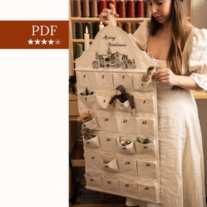 May include: A beige fabric advent calendar with pockets numbered 1-24. The top has a handle and the words "Merry Christmas" with a village design. Some pockets contain small decorative items.