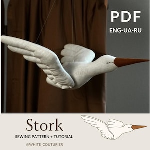 Puede incluir: Un patrón de costura y un tutorial para hacer un móvil de cigüeña blanca. El patrón incluye instrucciones en inglés, ucraniano y ruso. La imagen muestra un móvil de cigüeña terminado colgando de una cortina marrón.