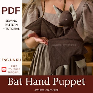 May include: A brown bat hand puppet with a light grey face and ears. The puppet has outstretched wings and is made of fabric. The image also includes text that says "PDF Sewing Pattern + Tutorial" and "Bat Hand Puppet".