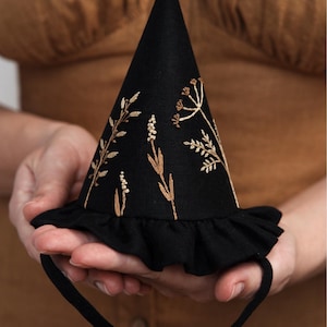 Puede incluir: Un sombrero de bruja negro con un borde rizado y un dise&ntilde;o de bordado floral marr&oacute;n. El sombrero tiene una cinta negra.