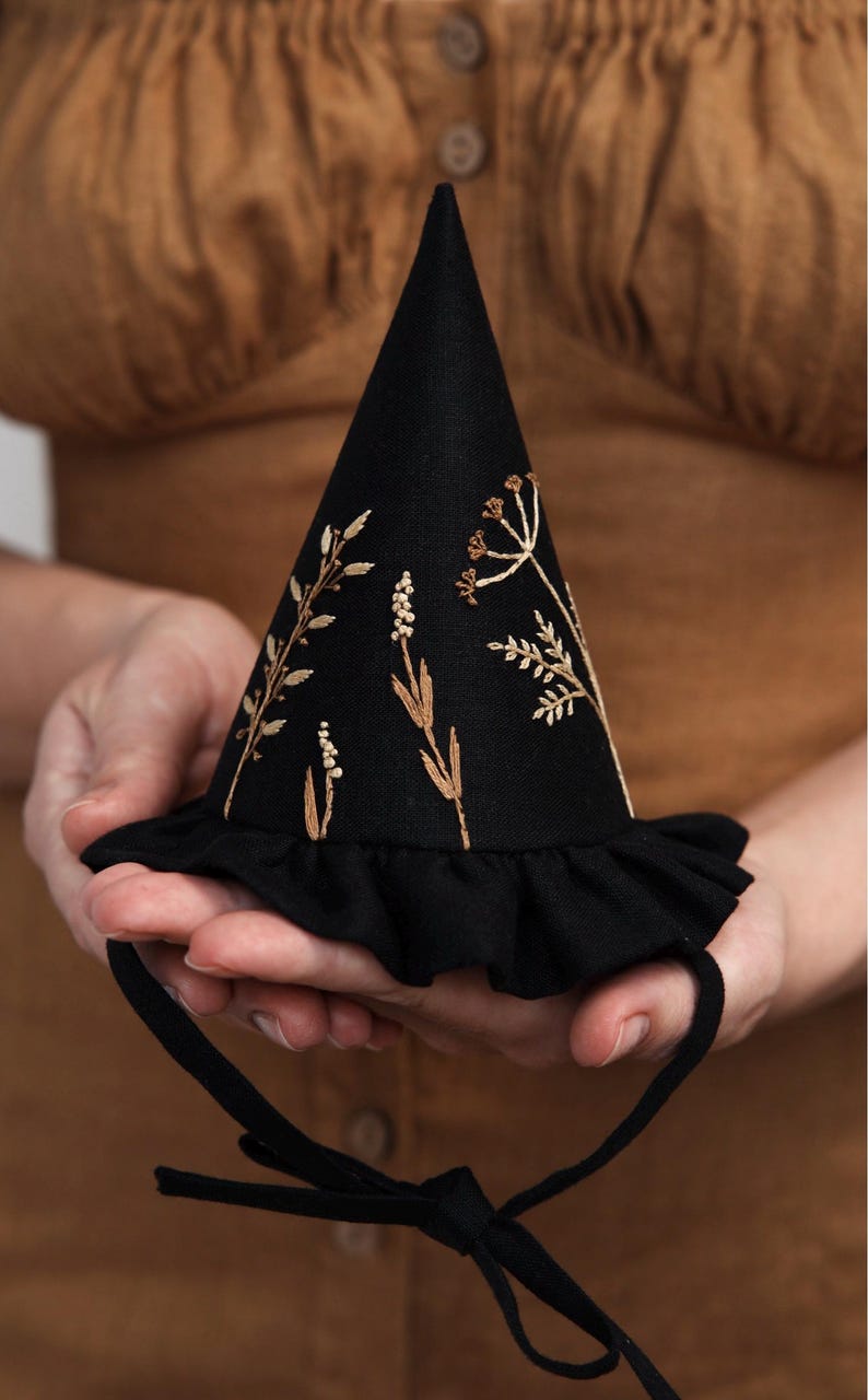 Puede incluir: Un sombrero de bruja negro con un borde rizado y un dise&ntilde;o de bordado floral marr&oacute;n. El sombrero tiene una cinta negra.