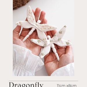 Op de afbeelding: Twee witte stoffen libellenornamenten, elk ongeveer 11 cm x 8 cm groot, worden in open handen gehouden. De vleugels zijn versierd met delicate goudkleurige borduursels. De tekst "Dragonfly" en "WHITE-COUTURIER.COM" zijn zichtbaar.