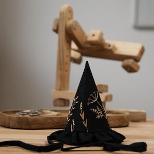 Puede incluir: Un sombrero negro en forma de cono con un dise&ntilde;o floral bordado en oro y un ribete de volantes negro. El sombrero est&aacute; sobre una superficie de madera.