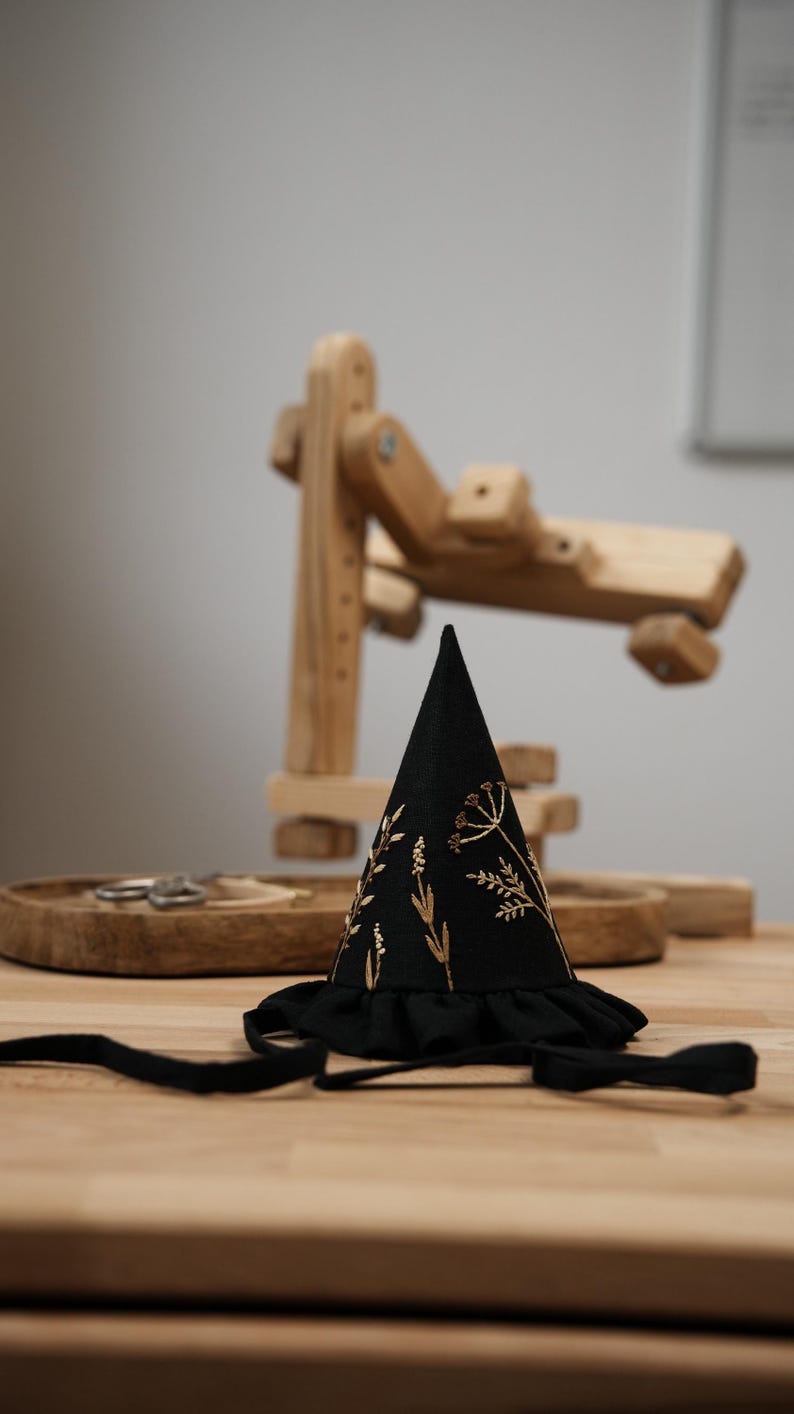 Puede incluir: Un sombrero negro en forma de cono con un dise&ntilde;o floral bordado en oro y un ribete de volantes negro. El sombrero est&aacute; sobre una superficie de madera.