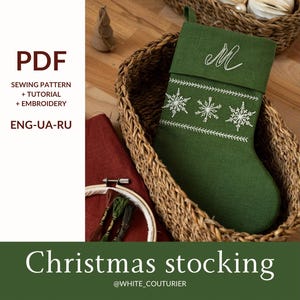 Puede incluir: Una media de Navidad verde con bordado de copos de nieve blancos y la letra "M" bordada en la parte superior. La media está en una cesta de mimbre con adornos de papel blanco.