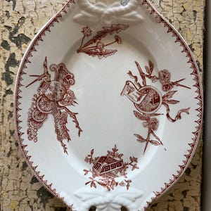 May include: An oval white ceramic serving dish featuring a dark red transferware pattern of floral motifs and geometric shapes. The dish has a scalloped rim with a decorative border and two integrated handles at the ends with embossed leaf details.