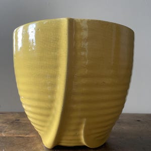 Könnte beinhalten: Ein gelber Keramik-Pflanztopf mit geripptem Design im unteren Bereich. Der Pflanztopf hat eine einzigartige Form mit leicht ausgestelltem Oberteil und einer strukturierten, rissigen Oberfläche. Der Pflanztopf steht auf einer Holzoberfläche.