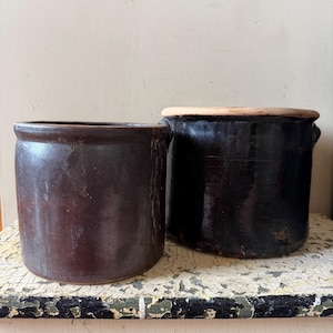 Pode incluir: Dois potes cilíndricos de cerâmica com acabamentos esmaltados em castanho escuro. O pote mais pequeno à esquerda tem uma superfície lisa e mate, enquanto o pote maior à direita apresenta um esmalte castanho escuro brilhante e uma tampa plana clara.