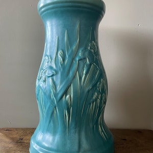 Puede incluir: Un pedestal de cerámica azul verdoso con un diseño en relieve de iris y hojas.