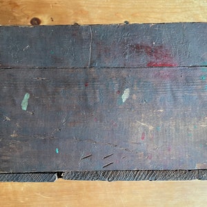May include: A wooden box with a dark brown finish. The box has a worn, rustic appearance with visible signs of age and use. There are several small paint splatters on the surface, including red, green, and blue.