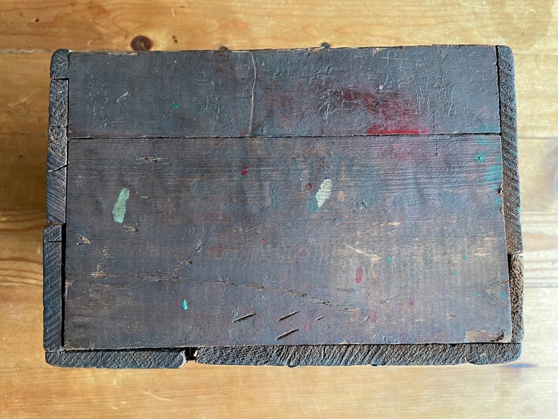 May include: A wooden box with a dark brown finish. The box has a worn, rustic appearance with visible signs of age and use. There are several small paint splatters on the surface, including red, green, and blue.