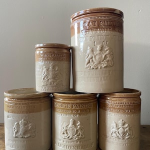 May include: A collection of antique stoneware jars in varying sizes. The jars are cream-colored with brown rims and embossed text and heraldic crests. The text includes "GEORGE PAINTER" and "PURVEYOR TO HRH THE PRINCE OF WALES".