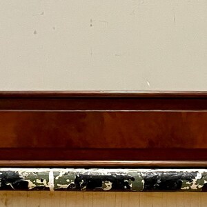 May include: A rectangular wooden box with a dark brown finish and a lighter wood inlay. The box sits on a distressed, painted surface, with a neutral background. The box appears to be a vintage or antique item.