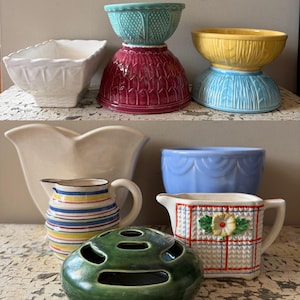 Könnte beinhalten: Eine Sammlung von Vintage-Keramikschalen, -krügen und Dekorationsartikeln in verschiedenen Farben. Enthält eine weiße quadratische Schale, eine türkisfarbene Schale auf einem weinroten Sockel, eine gelbe Schale auf einem blauen Sockel, einen weißen Krug, einen gestreiften Krug, eine blaue Schale, einen grünen Blumenfrosch und einen kleinen Krug mit Blumendesign.