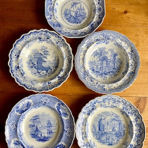 May include: Five antique blue and white porcelain plates with scalloped edges. Each plate features a different scenic landscape design in the center, surrounded by floral and geometric patterns. The plates are arranged on a wooden surface.