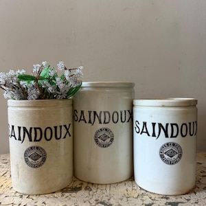 May include: Three cylindrical ceramic crocks with off-white and cream finishes. Each container features the black text SAINDOUX and a circular Felix Potin Paris logo. The leftmost crock contains a decorative arrangement of small beaded flowers.