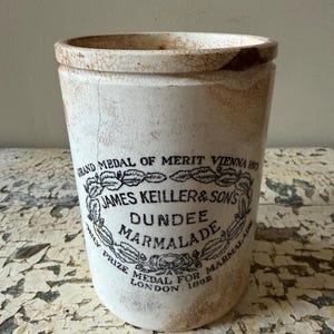 May include: Antique white ceramic jar with a slightly flared shape. The jar features a black and white label with the text "JAMES KEILLER & SONS DUNDEE MARMALADE" and "GRAND MEDAL OF MERIT VIENNA 1873". The jar has a distressed, aged appearance.