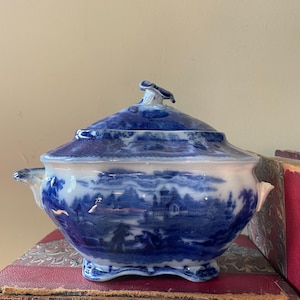 May include: A blue and white ceramic serving dish with a lid. The dish features a landscape design and has handles on either side. The lid has a decorative finial. The dish is sitting on a red book.