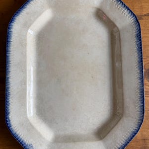 Könnte beinhalten: Eine achteckige Servierplatte aus Keramik mit cremefarbener Mitte und einem dunkelblauen, federartigen Randmuster. Die Platte verfügt über einen breiten, flachen Rand mit einem texturierten, gerippten Design, das das glatte, leicht vertiefte Becken einrahmt.
