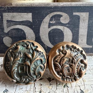 May include: Two antique brass cabinet knobs with ornate floral designs. One knob has a verdigris patina, while the other is a polished brass color. The knobs are round and have a raised relief pattern. The background includes a distressed wooden sign with the number 561.
