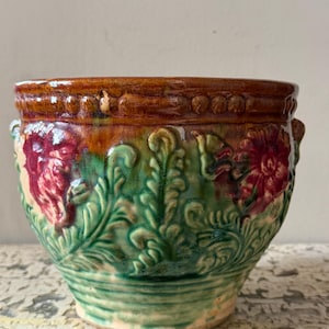 May include: A round ceramic planter featuring a glossy brown rim with a beaded texture. The body displays a relief pattern of green foliage and red flowers. The base is a light tan colour with horizontal ridges, and the entire piece has a glazed finish.