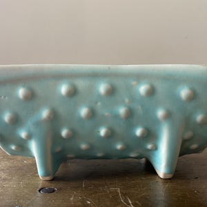 Pode incluir: Um vaso retangular de cerâmica azul claro com uma superfície texturizada de pequenas protuberâncias arredondadas brancas. O vaso assenta em três pernas curtas com pontas brancas. Esta peça decorativa é adequada para pequenas plantas ou suculentas.