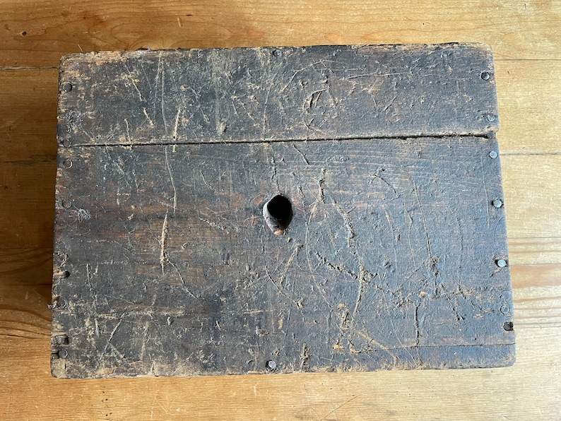 May include: A dark brown wooden box with a single hole in the top. The box is made of rough-hewn wood and has a worn, rustic appearance.