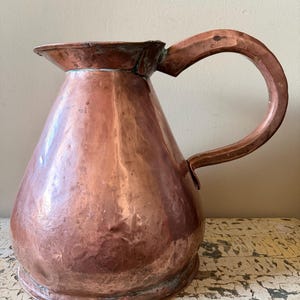 May include: A copper pitcher with a wide base and a curved handle. The pitcher has a spout and a slightly flared rim. The copper has a warm, reddish-brown colour with some areas of patina, suggesting age and use. The handle is securely attached to the body.